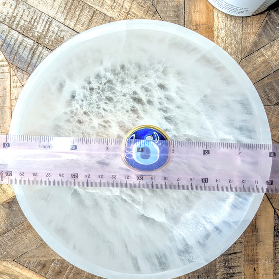 A round extra large selenite bowl, fibrous white surface, shown on a rustic wood background with a pink measuring tape across the center and a small blue and gold circular item placed in the middle.