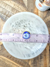 A round extra large selenite bowl, fibrous white surface, shown on a rustic wood background with a pink measuring tape across the center and a small blue and gold circular item placed in the middle.