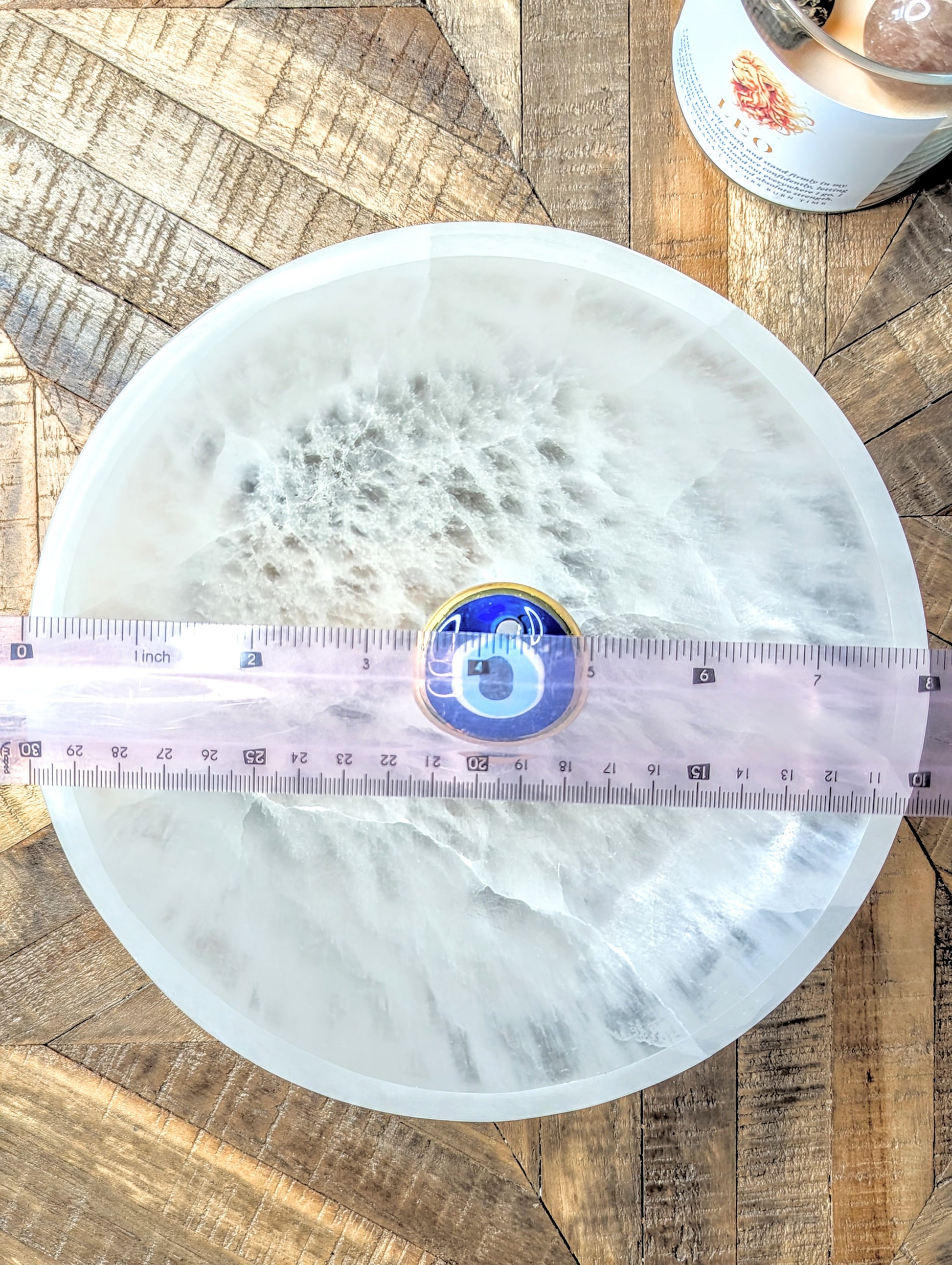 A round extra large selenite bowl, fibrous white surface, shown on a rustic wood background with a pink measuring tape across the center and a small blue and gold circular item placed in the middle.