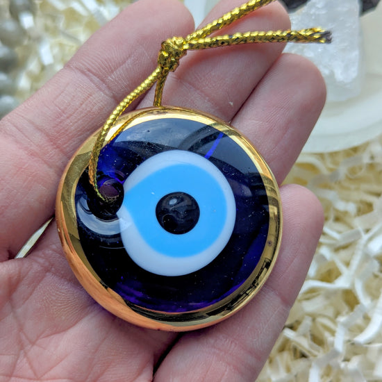 A hand holding a round glass evil eye pendant with a gold border, featuring blue, white, and black concentric circles, complete with a gold hanging cord.