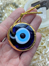 A hand holding a round glass evil eye pendant with a gold border, featuring blue, white, and black concentric circles, complete with a gold hanging cord.