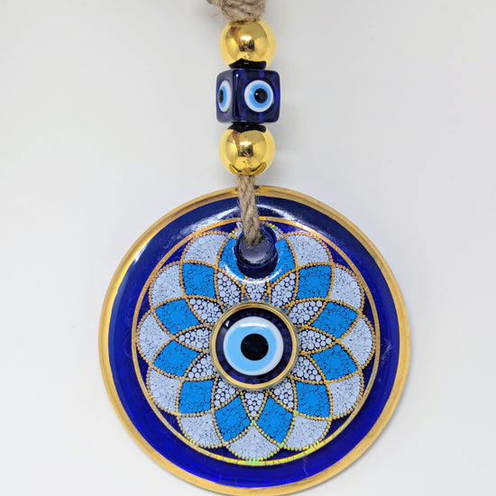 A circular evil eye (Nazar) wall hanging ornament with a blue and white geometric pattern, gold trim, and a beige jute rope with decorative blue and gold beads at the top, shown on a plain white background.