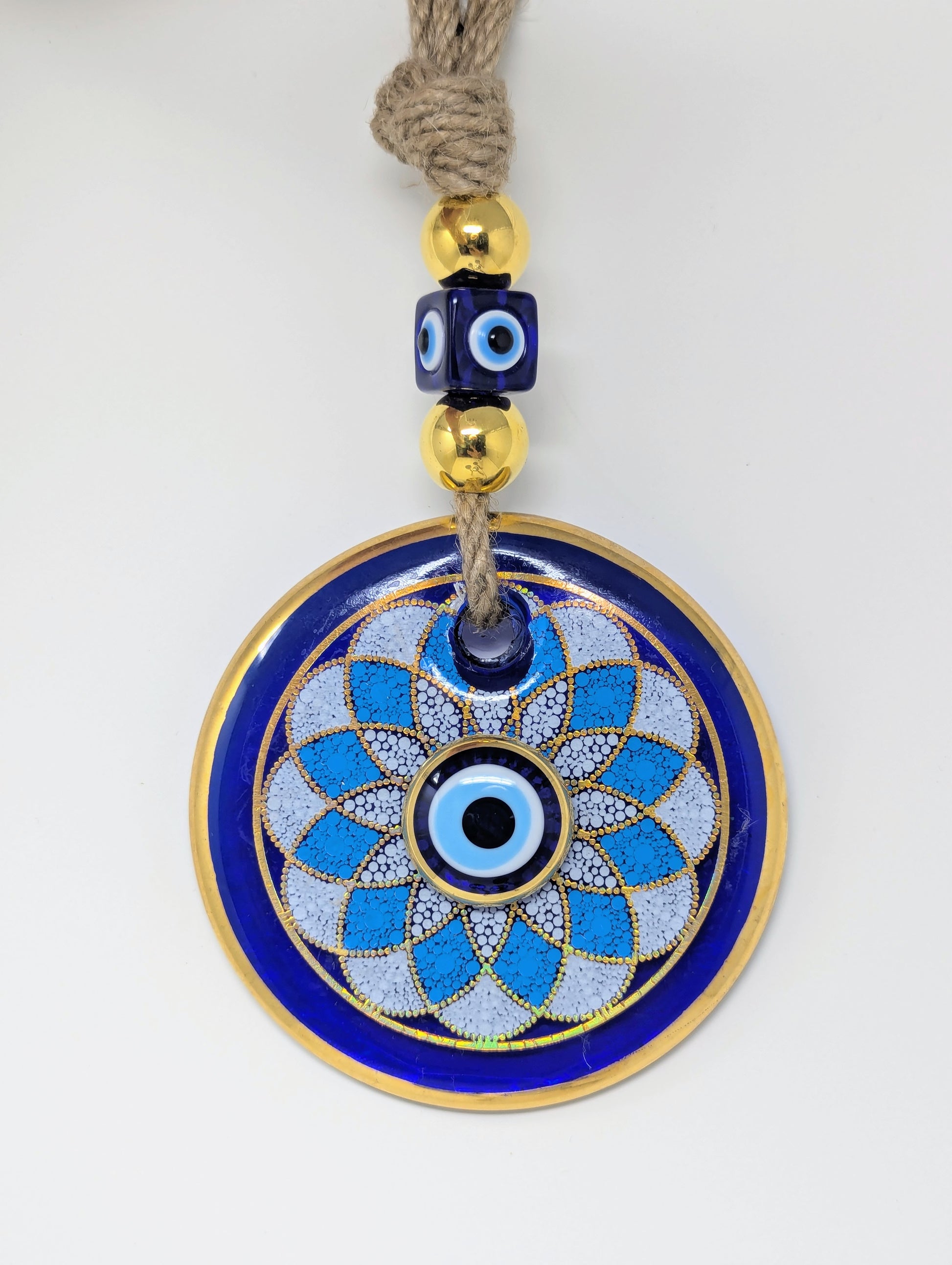 A circular evil eye (Nazar) wall hanging ornament with a blue and white geometric pattern, gold trim, and a beige jute rope with decorative blue and gold beads at the top, shown on a plain white background.