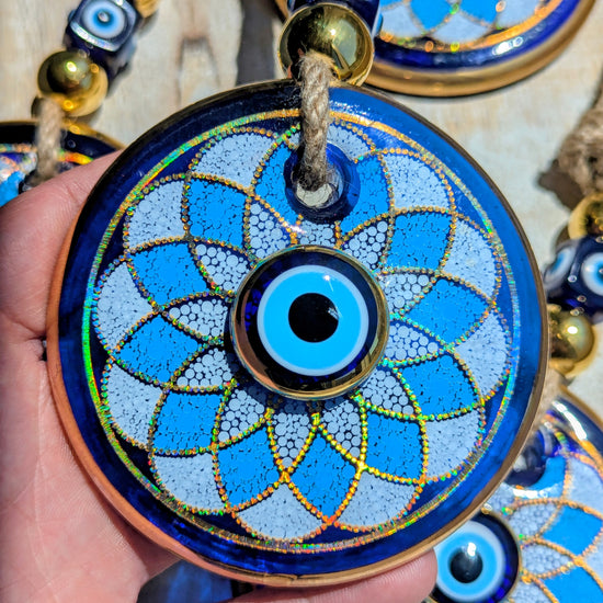 A hand holds a round blue and white evil eye pendant ornament with gold detailing and a rope loop, with other similar pendants visible in the background.