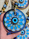 A hand holds a round blue and white evil eye pendant ornament with gold detailing and a rope loop, with other similar pendants visible in the background.