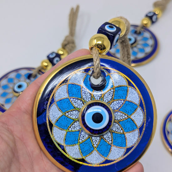 A hand holds a round blue and white evil eye pendant with gold trim and a tassel, with other similar pendants blurred in the background.