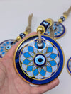 A hand holds a round blue and white evil eye pendant with gold trim and a tassel, with other similar pendants blurred in the background.