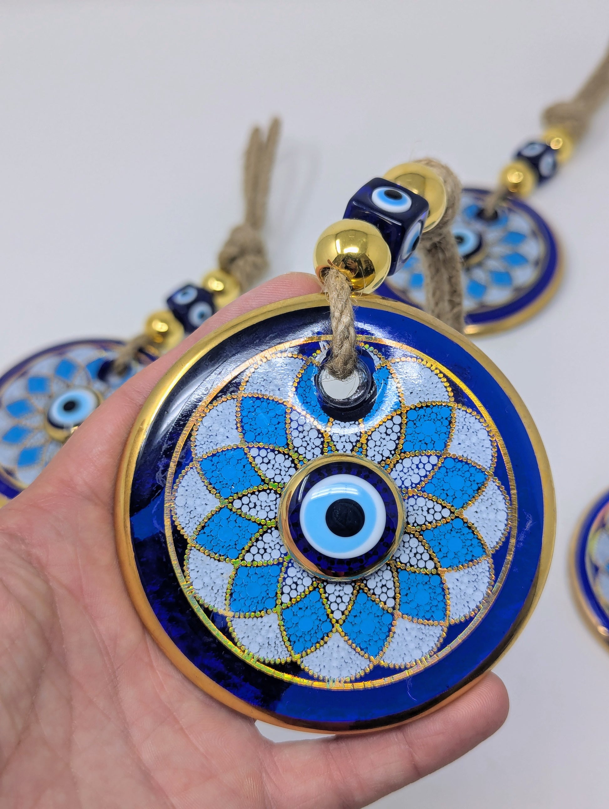 A hand holds a round blue and white evil eye pendant with gold trim and a tassel, with other similar pendants blurred in the background.