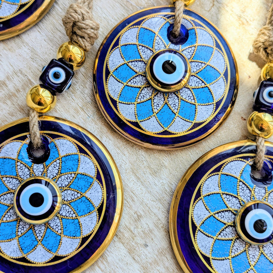 Close-up of several round evil eye hanging ornaments with blue enamel, gold trim, white textured centers, and knotted cords on a light wood surface.