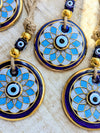 Close-up of several round evil eye hanging ornaments with blue enamel, gold trim, white textured centers, and knotted cords on a light wood surface.