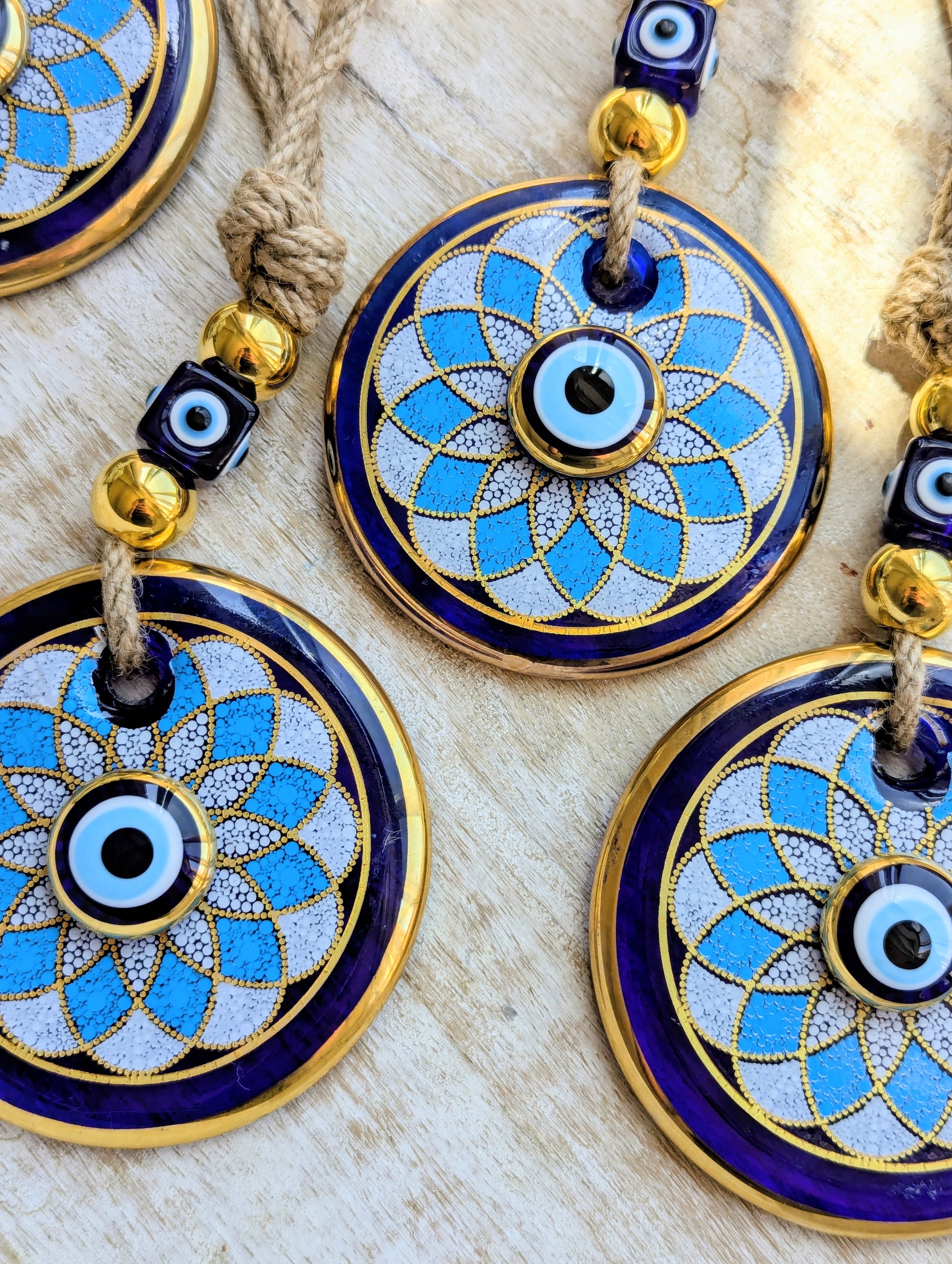 Close-up of several round evil eye hanging ornaments with blue enamel, gold trim, white textured centers, and knotted cords on a light wood surface.