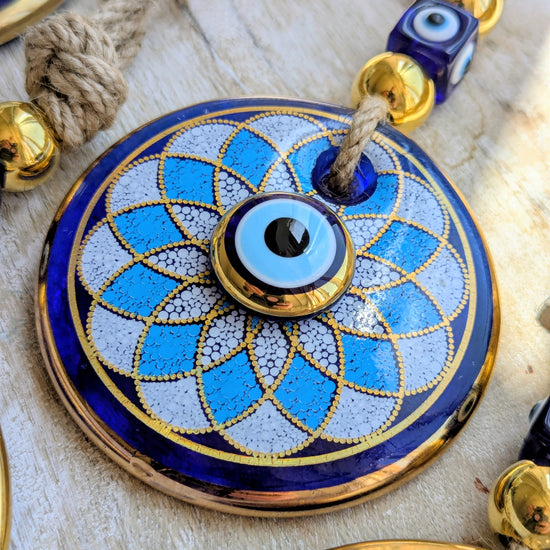 A close-up of a round blue and white evil eye pendant necklace with gold trim, hanging on a rope with blue and gold beads.