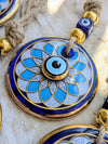 A close-up of a round blue and white evil eye pendant necklace with gold trim, hanging on a rope with blue and gold beads.