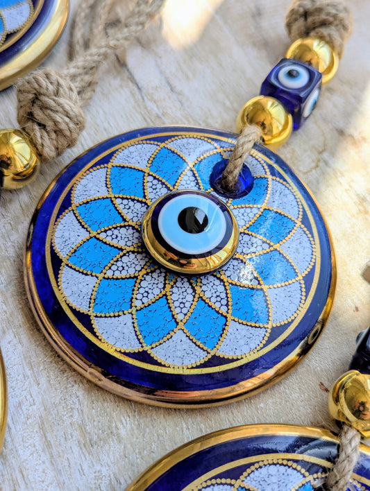 A close-up of a round blue and white evil eye pendant necklace with gold trim, hanging on a rope with blue and gold beads.