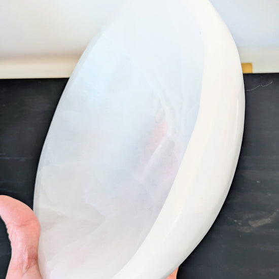 A hand holding a large, smooth, white selenite bowl or plaque against a dark tabletop and light wall background.