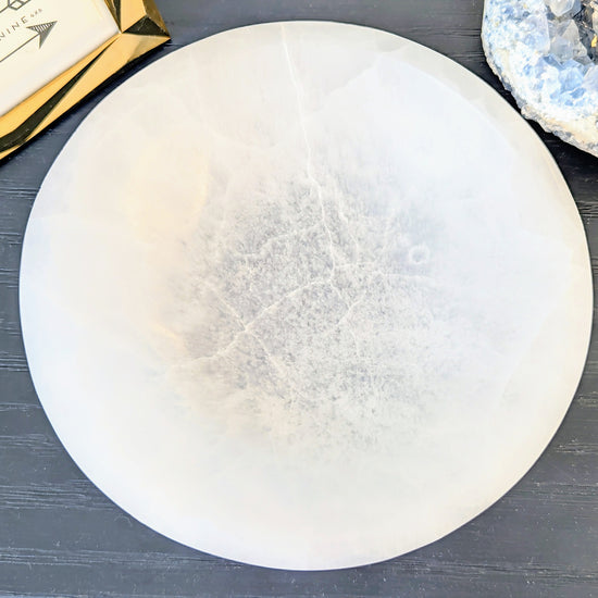 A large white round selenite bowl, displayed on a dark gray wood surface next to a gold box and a blue geode specimen.