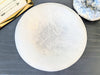 A large white round selenite bowl, displayed on a dark gray wood surface next to a gold box and a blue geode specimen.