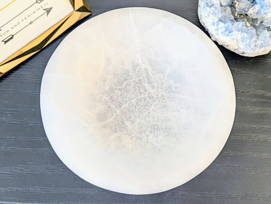 A large white round selenite bowl, displayed on a dark gray wood surface next to a gold box and a blue geode specimen.