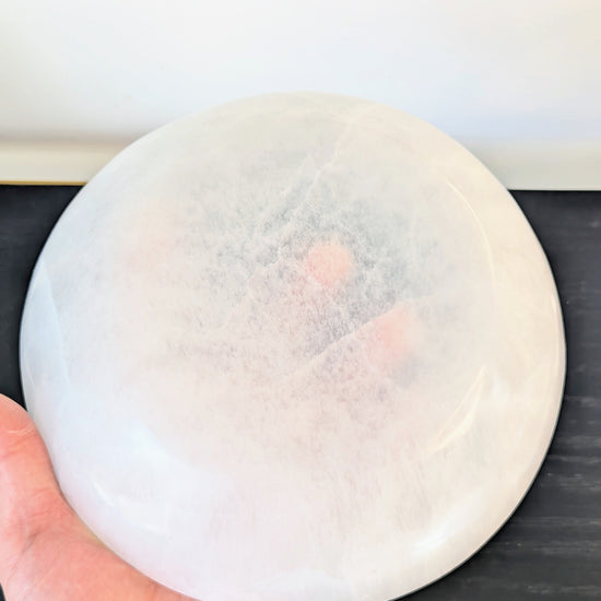 A hand holding a large, round, translucent white selenite crystal bowl against a dark tabletop and light wall background.