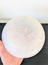 A hand holding a large, round, translucent white selenite crystal bowl against a dark tabletop and light wall background.