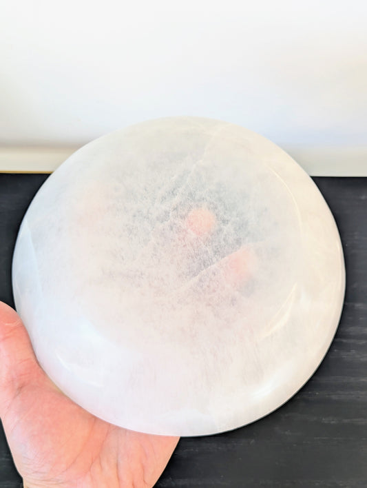 A hand holding a large, round, translucent white selenite crystal bowl against a dark tabletop and light wall background.