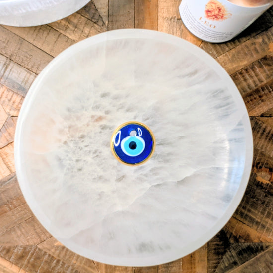 A round white extra large selenite crystal bowl with a central blue and gold evil eye charm, photographed from above on a rustic wooden surface with other decorative items partially visible.