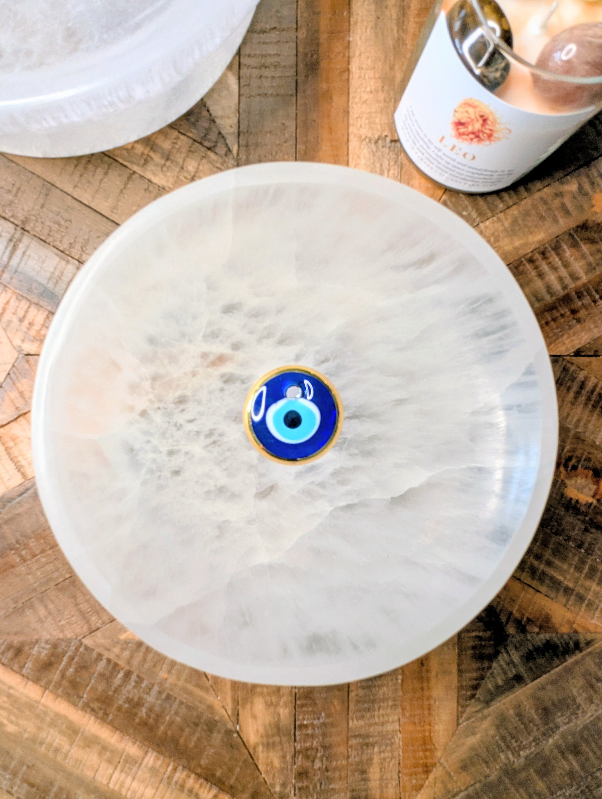 A round white extra large selenite crystal bowl with a central blue and gold evil eye charm, photographed from above on a rustic wooden surface with other decorative items partially visible.