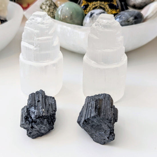 Two white selenite towers with two black magnetite clusters in front, displayed on a white surface with other crystals and a bowl in the background.