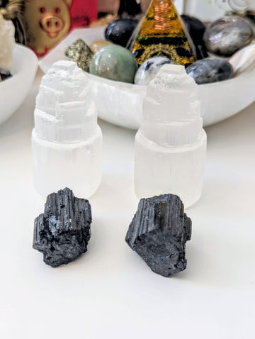 Two white selenite towers with two black magnetite clusters in front, displayed on a white surface with other crystals and a bowl in the background.