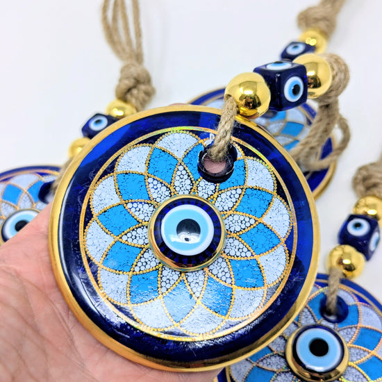 A hand holds a round blue and gold evil eye pendant ornament with a woven rope tassel, with additional similar ornaments partially visible in the background against a plain white backdrop.