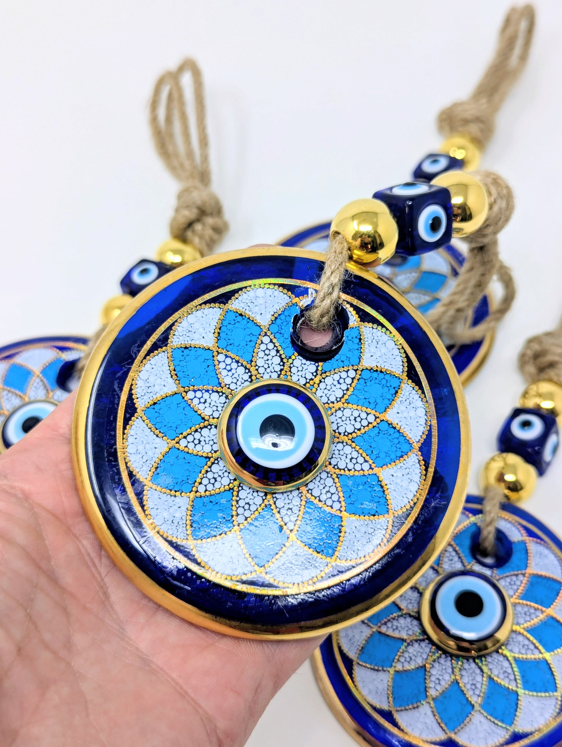 A hand holds a round blue and gold evil eye pendant ornament with a woven rope tassel, with additional similar ornaments partially visible in the background against a plain white backdrop.