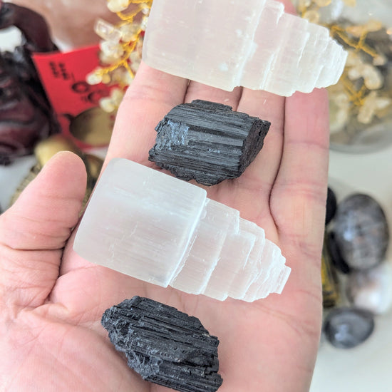 A hand holds three crystal clusters: two milky white selenite stairpyramids and one dark charcoal black tourmaline geode, with blurred crystals in the background.