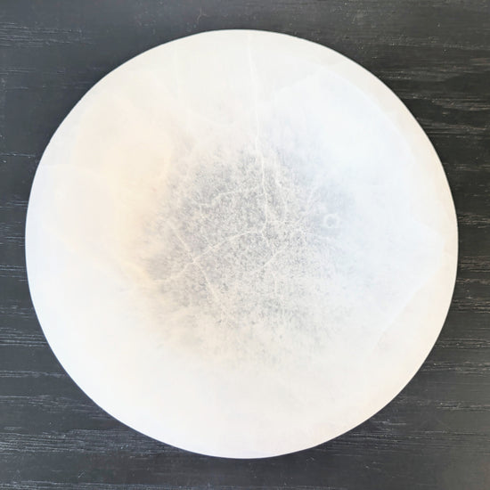 A round selenite bowl with a textured, cloudy interior and fine crack-like lines, displayed on a dark wood background.