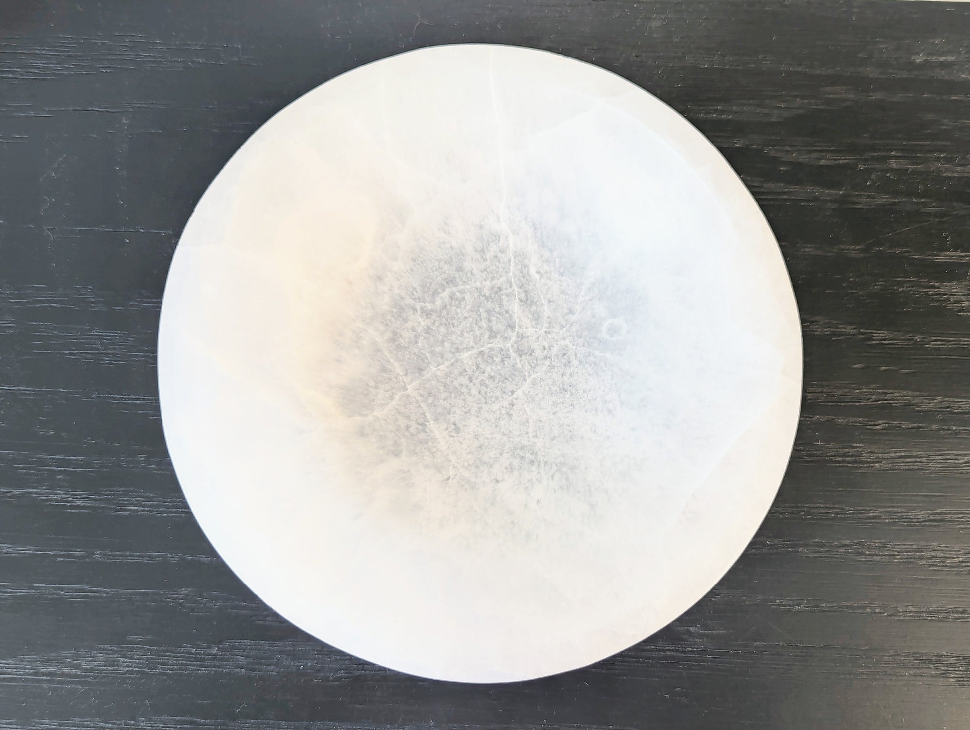 A round selenite bowl with a textured, cloudy interior and fine crack-like lines, displayed on a dark wood background.