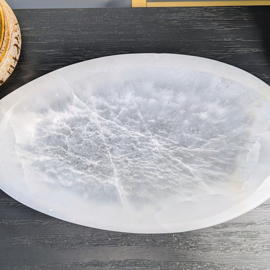 An oval extra large selenite bowl with natural crystalline textures, displayed on a dark gray wood surface.