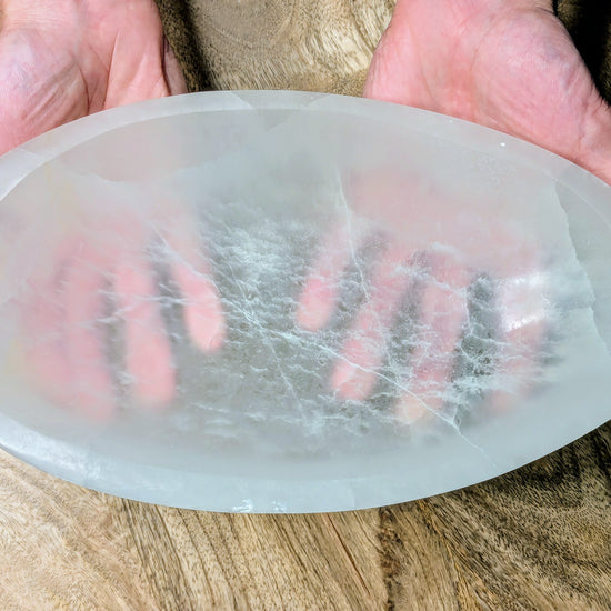 A large, oval-shaped translucent white selenite bowl visible internal fibrous structures, held in two hands over a wooden surface.