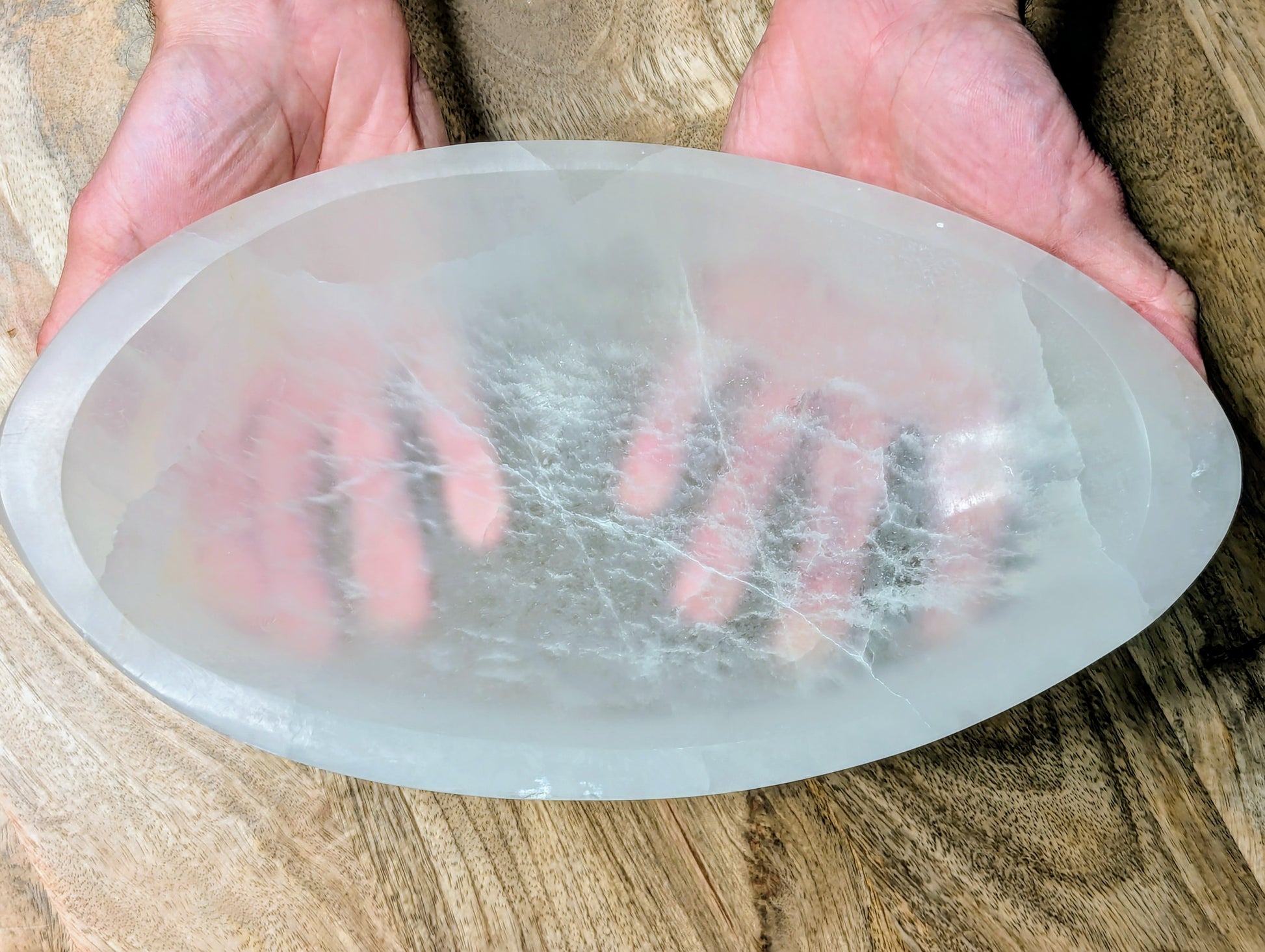 A large, oval-shaped translucent white selenite bowl visible internal fibrous structures, held in two hands over a wooden surface.