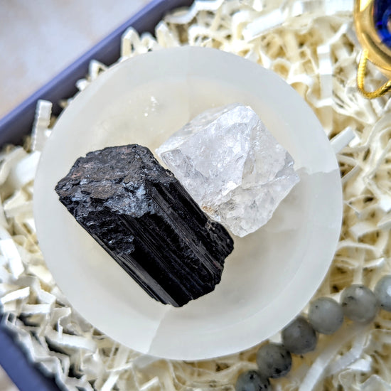 A pair of raw mineral crystals, one black and one clear, displayed on a white circular stand inside a gift box filled with white paper shred.