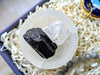 A pair of raw mineral crystals, one black and one clear, displayed on a white circular stand inside a gift box filled with white paper shred.
