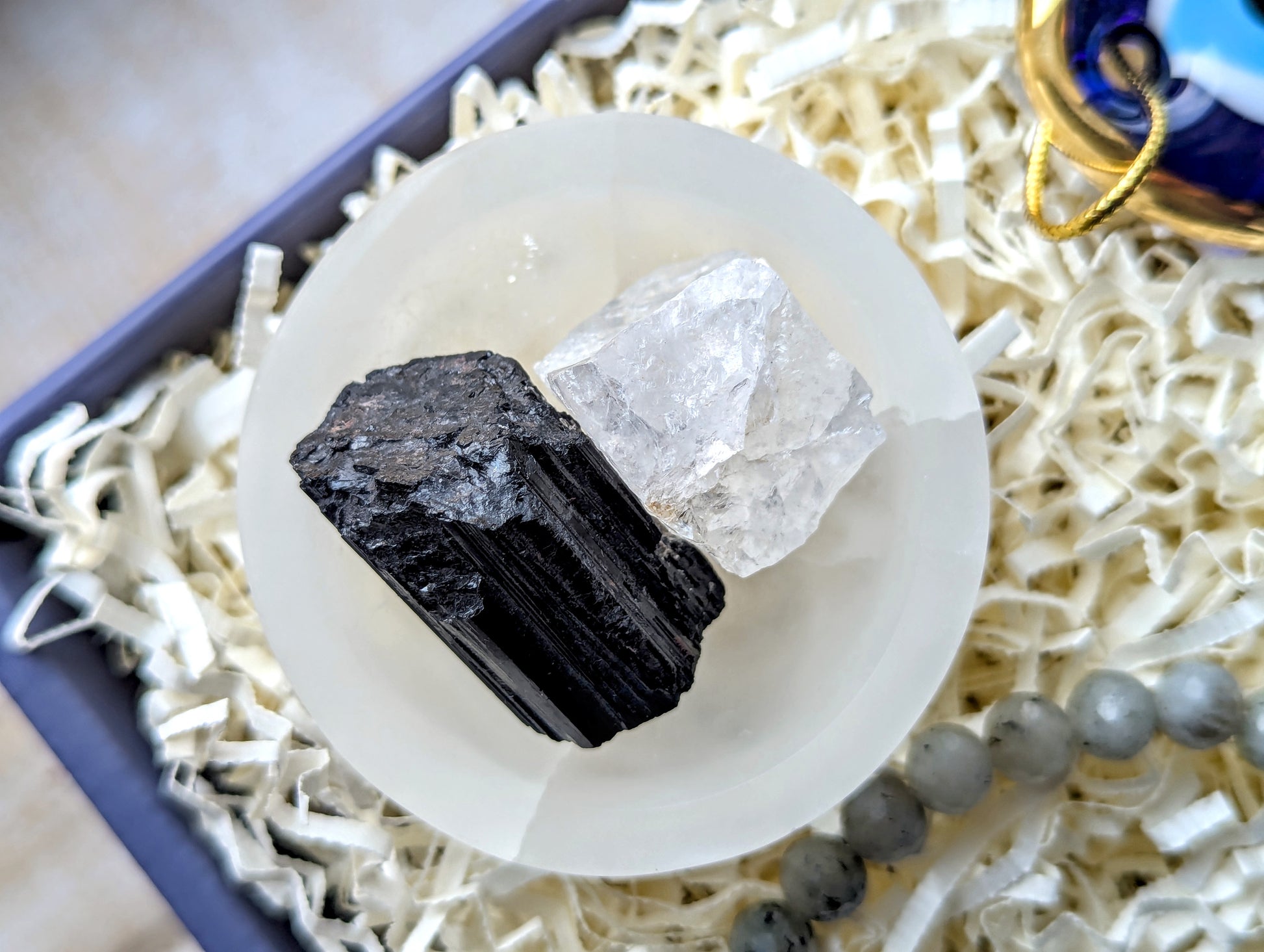 A pair of raw mineral crystals, one black and one clear, displayed on a white circular stand inside a gift box filled with white paper shred.