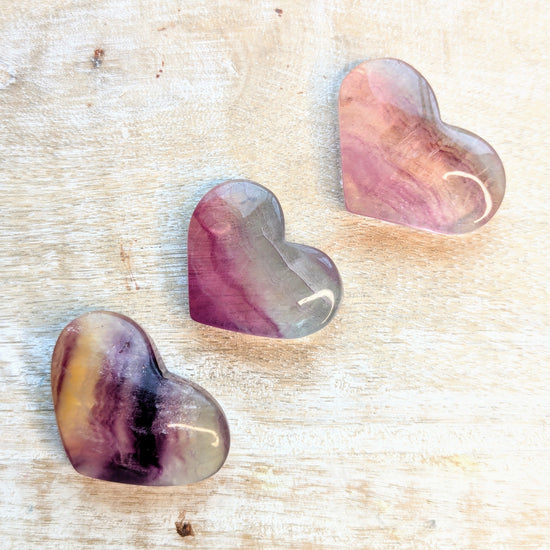 Three heart-shaped rainbow fluorite stones with purple and pink hues on a textured beige surface