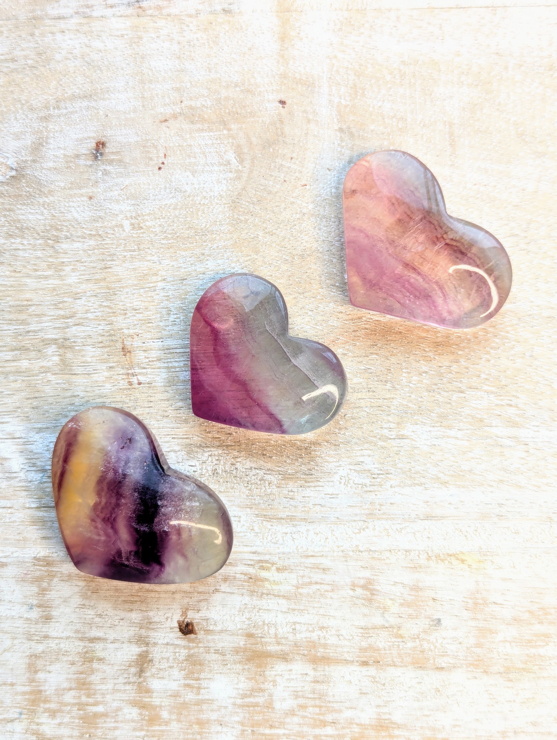 Three heart-shaped rainbow fluorite stones with purple and pink hues on a textured beige surface