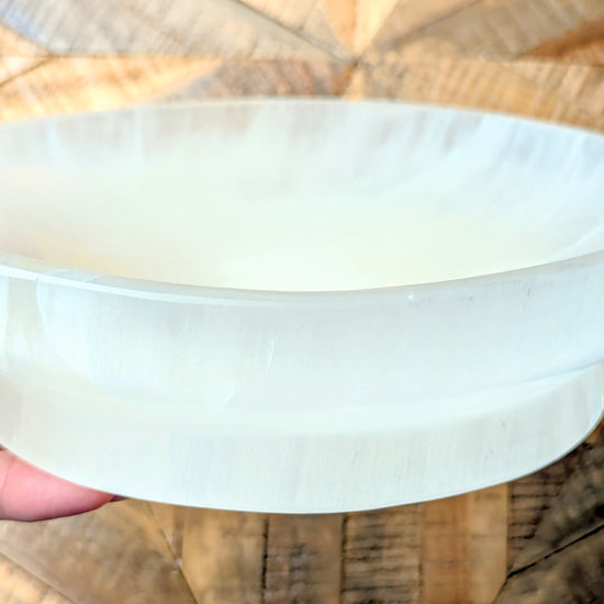 A hand holding a large, round, off-white translucent selenite bowl or dish against a wooden background.