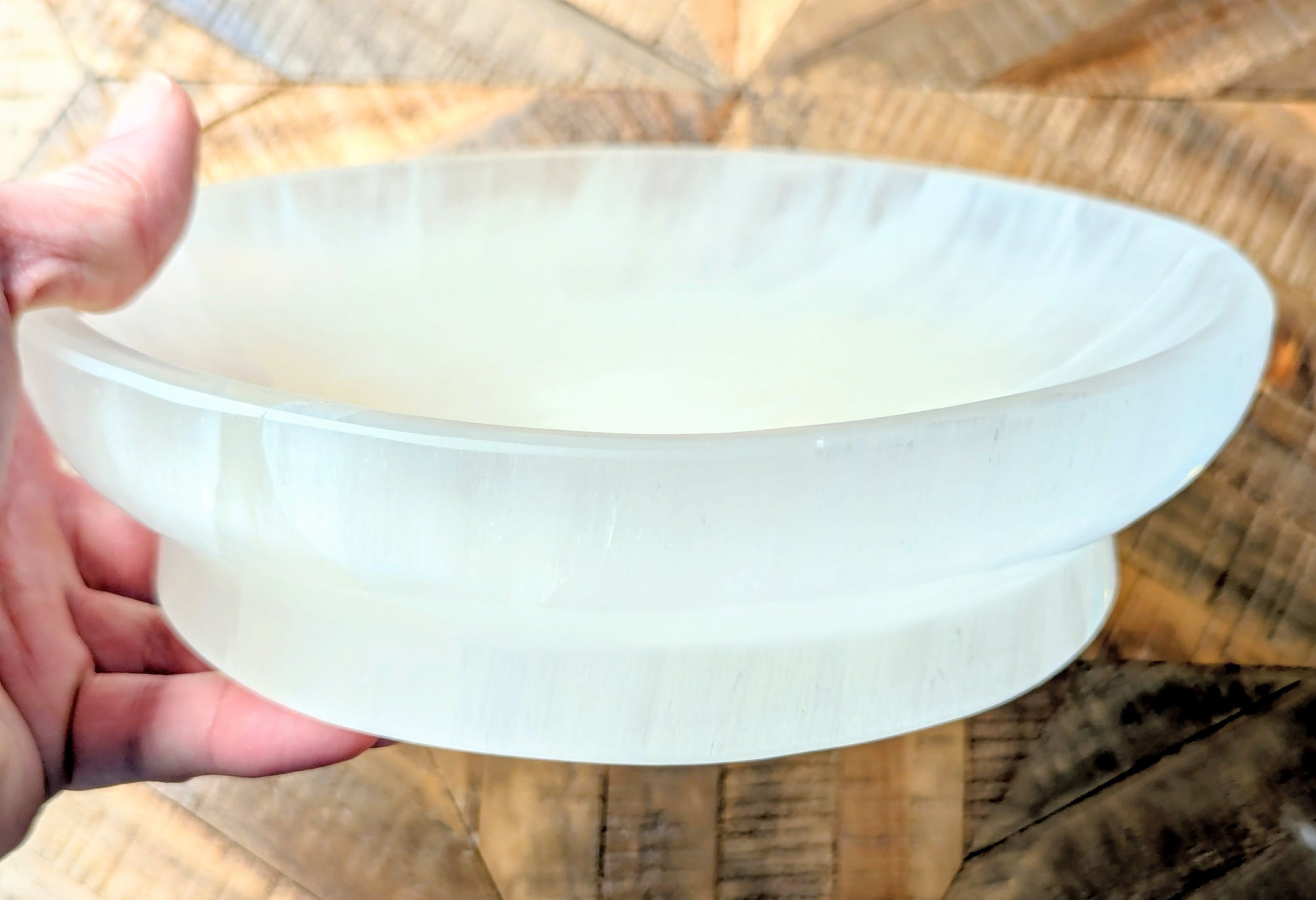 A hand holding a large, round, off-white translucent selenite bowl or dish against a wooden background.
