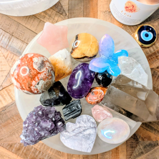 A collection of colorful raw crystals and gemstones arranged on a selenite bowl including a purple amethyst heart, a pink star, a blue butterfly, and various polished stones, set on a wooden table next to a candle.