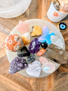 A collection of colorful raw crystals and gemstones arranged on a selenite bowl including a purple amethyst heart, a pink star, a blue butterfly, and various polished stones, set on a wooden table next to a candle.