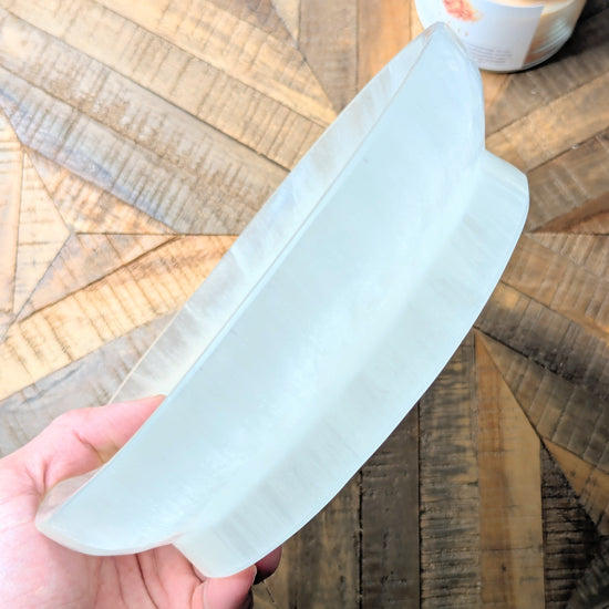 A hand holding a thick, translucent white selenite bowl over a geometric wood-patterned surface.