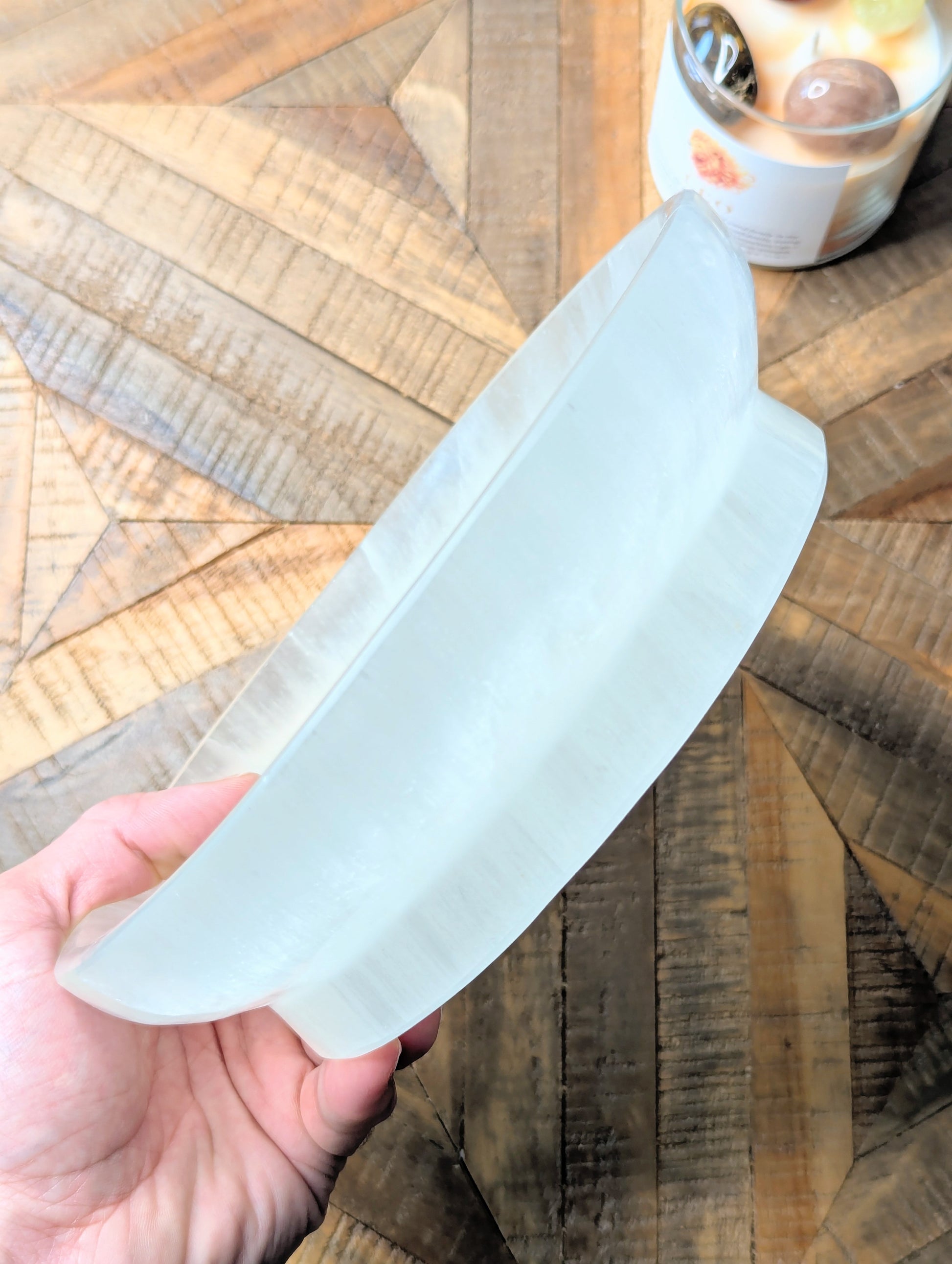 A hand holding a thick, translucent white selenite bowl over a geometric wood-patterned surface.
