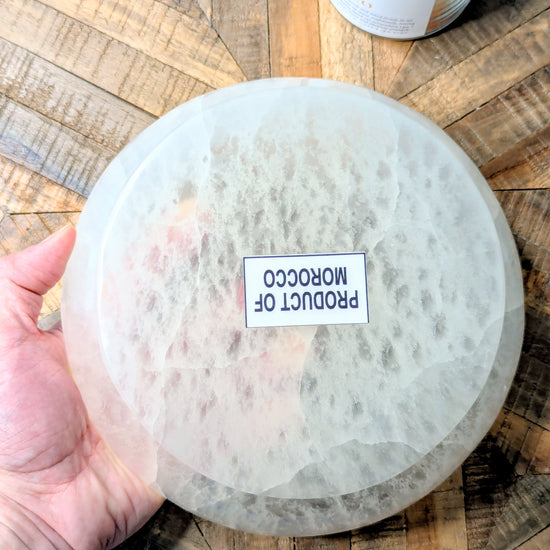 A hand holds a large, round, translucent white selenite bowl with a textured surface. A small white label on the front reads 'PRODUCT OF MOROCCO'.