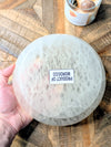 A hand holds a large, round, translucent white selenite bowl with a textured surface. A small white label on the front reads 'PRODUCT OF MOROCCO'.